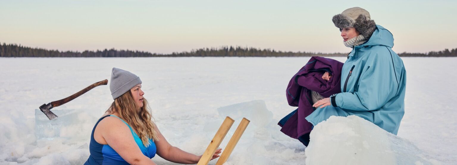 Två kvinnor vinterbadar i isvak på vacker sjö